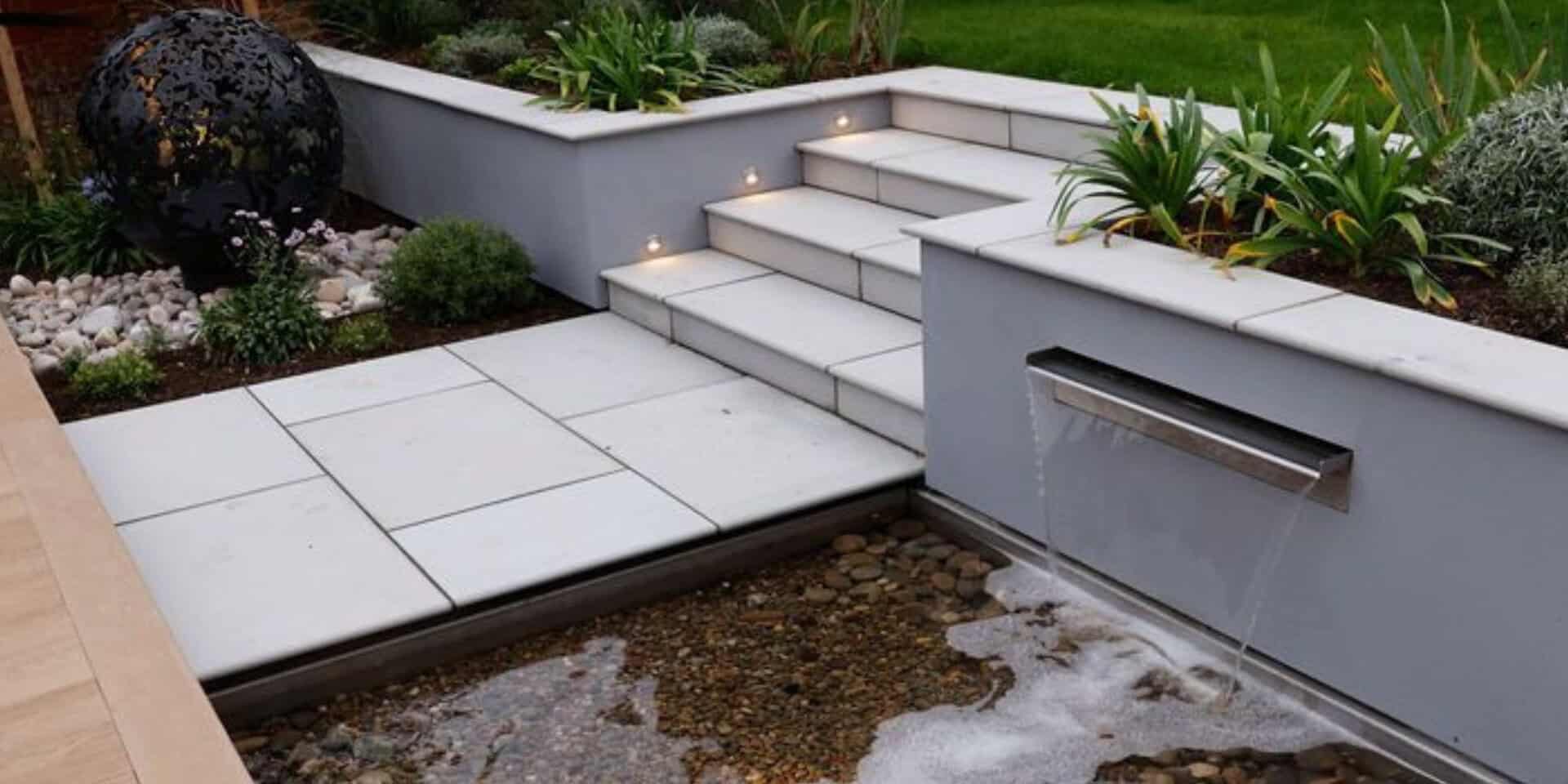 Garden water blade feature which creates a sheet of water tumbling into a small pool. A beautiful garden focal point surrounded by planting and next to a porcelain paved path.